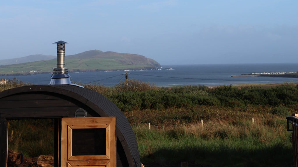 A scenic landscape photograph featuring a vast blue sky with wispy white clouds, a distant green hill, and the sea. In the foreground, the top portion of a dark wooden cabana or hut is visible on a grassy shore.