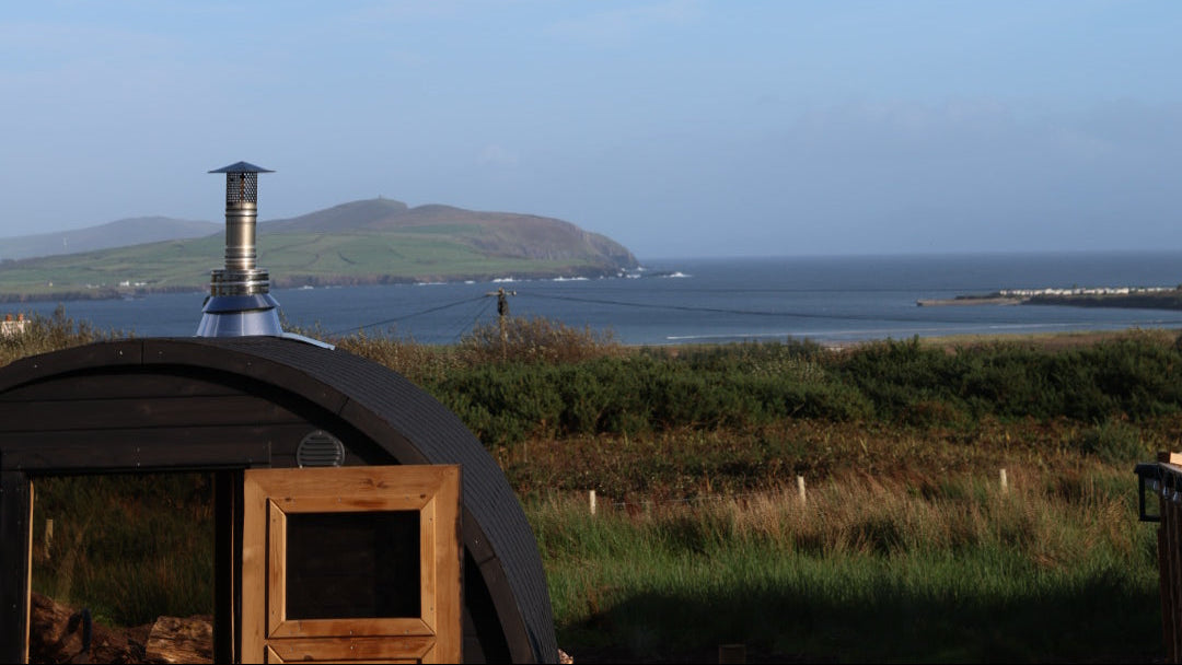 A scenic landscape photograph featuring a vast blue sky with wispy white clouds, a distant green hill, and the sea. In the foreground, the top portion of a dark wooden cabana or hut is visible on a grassy shore.
